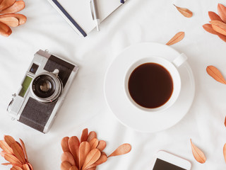 top view of coffee shop concept with coffee cup, notebook, dry leaf, retro camera and smartphone on white fabric background.