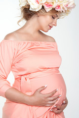Beautiful pregnant woman in a silk pink dress and a wreath of flowers on her head. Studio, white background.
