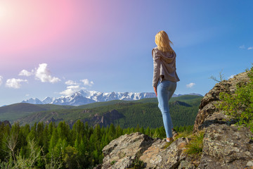 Naklejka premium Young blonde girl stay on a rock on a sunny warm autumn summer day resting while traveling near a green tree on the background of landscape snow-capped mountain top and a blue sky with clouds in Altai