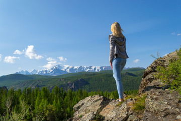 Young pretty woman stay on a rock on a sunny warm autumn summer day resting while traveling near a green tree on the background of landscape snow-capped mountain top and a blue sky with clouds