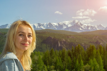 Portrait of a young beautiful blonde who smiles and looks into the camera against the backdrop of the large snow-covered Altai mountains on a clear summer day