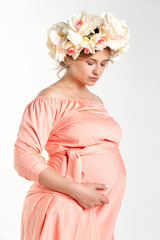 Beautiful pregnant woman in a silk pink dress and a wreath of flowers on her head. Studio, white background.
