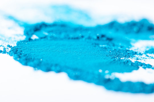 Bright blue eyeshadow crushed and smeared against a white background.  Shallow depth of field.  Horizontal image.