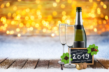 2019 Silvester bokeh hintergund mit Sektflasche Sektgläsern Tafel mit Grüßen und Wünschen im Schnee auf Holzboden