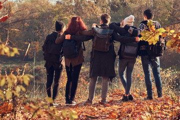 Back view of young friends hugging together and looking at the lake in beautiful autumn forest.
