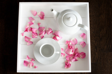 Weißes Teeservice aus Porzellan mit weißer Teekanne und weißer Tasse mit Untertasse auf weißem Tablett mit Rosenblättern dekoriert von oben