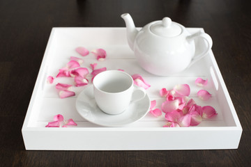 Weißes Teeservice aus Porzellan mit weißer Teekanne und weißer Tasse mit Untertasse auf weißem Tablett mit Rosenblättern dekoriert