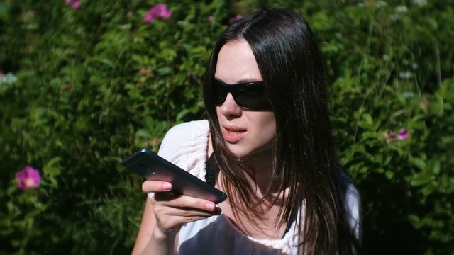 Beautiful brunette woman records a voice message on her mobile phone while sitting in the park on a Sunny day.