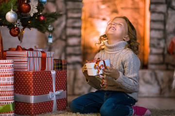Little girl child at home by the fireplace and Christmas tree with gifts and luminous garlands...