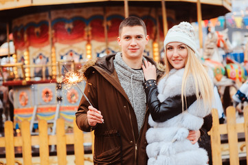 happy couple with christmas firelights walking in city holiday amusement park, carousel on background