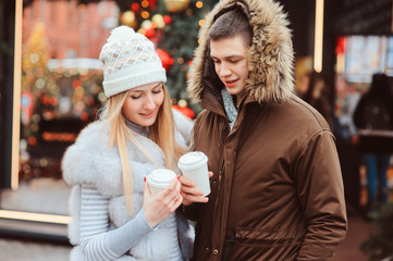 christmas portrait of happy couple with hot mulled wine or tea walking on city streets decorated for holidays, christmas trees with lights and toys on background