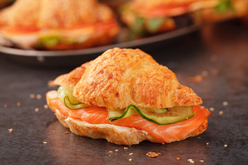Croissant sandwich with smoked salmon, cream cheese and cucumber slices on grey stone surface.