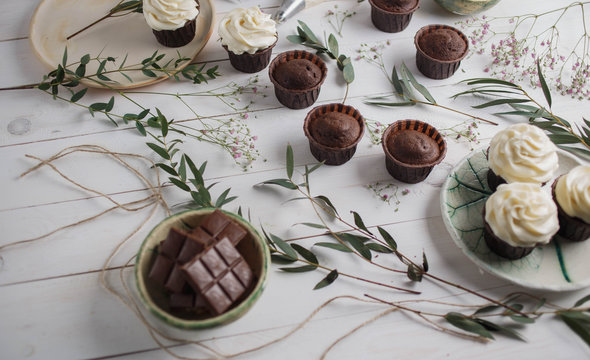 food photo, chocolate muffins and cupcakes white cream in ceramic plates of handwork on a white wooden background with green eucalyptus and baby's breath