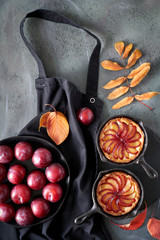 Red plums and plum tarts in cast iron skillets on a black cooking apron thrown on gray