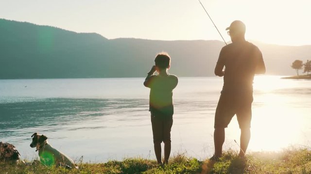 Slow motion slide dad and son, stand on the shore of a large and calm lake and fish with a fishing rod. A man and a boy cast spinning into a calm water surface. Lens flare. Dog Jack Russell Terrier