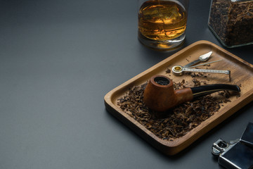 a smoking pipe and pipe tamper tool in wooden tray, a chrome lighter, tobacco jar with a glass of bourbon whiskey on the black table