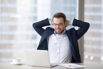Man sitting at table near laptop in office and feeling success or satisfaction. Boss or employee holds his arms behind head, guy leans back on chair, looks at computer screen and smiles in city office