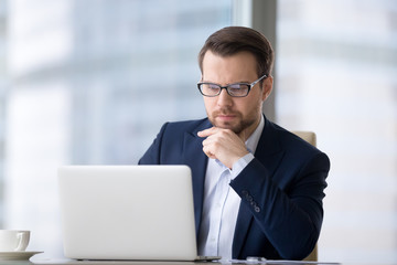 Fototapeta premium Caucasian man sitting at the table near laptop in the office and looking at the screen. Serious and pensive boss or employee working at his workplace and feeling interest or analyzing results.