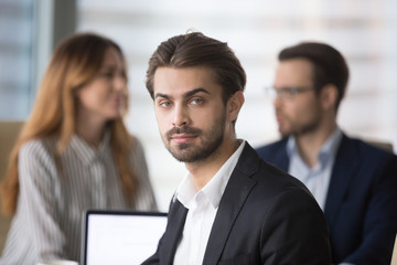 Obraz premium Head shot portrait of confident businessman or business owner at workplace, bearded man sitting at briefing and looking at camera. Colleagues discuss in background.