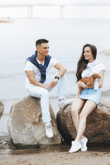 Young beautiful positive couple with small dog sitting on stone by sea