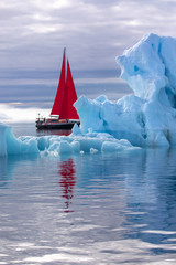 Beautiful red sailboat in the arctic next to a massive iceberg showing the scale. Ilulissat, Disko...