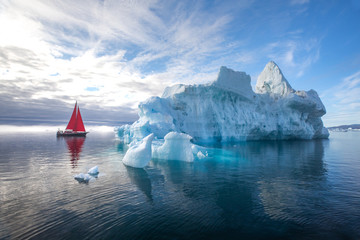 Beautiful red sailboat in the arctic next to a massive iceberg showing the scale. Ilulissat, Disko Bay, Greenland. - obrazy, fototapety, plakaty