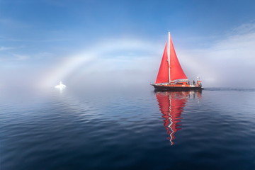 Beautiful red sailboat under a rainbow - fog bow on a foggy day lighted by sunlight. Disko bay,...