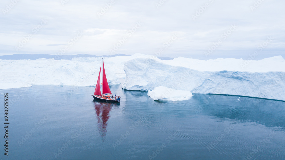 Wall mural beautiful red sailboat in the arctic next to a massive iceberg showing the scale - aerial view as vi