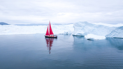 Beautiful red sailboat in the arctic next to a massive iceberg showing the scale -  aerial view as viewed from a drone. Ilulissat, Disko Bay, Greenland. © Kertu