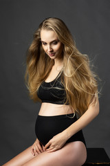 Fototapeta na wymiar A beautiful pregnant leggy blonde woman wearing black maternity underwear is sitting on a chair against a gray background. Health, fashion, motherhood. Advertising and commercial design.