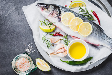Fresh fish sea bass and ingredients for cooking. Raw fish sea bass with spices and herbs on black slate table copy space.