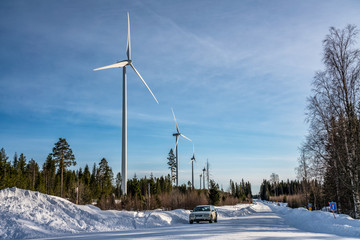 photo of windmill farm and car at straight road in winter forest, blue sky, sunny