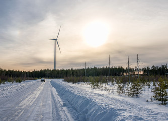photo of windmill, car at straight road and few dead pine trees in winter forest, blue sky, sunny