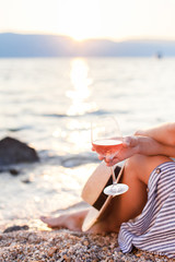 Girl is sitting on sea beach with wineglass of wine at sunset in summer vacation in resort. Tourist woman in striped dress with straw hat is enjoying life, view, relaxing, drinking, traveling.