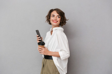 Fototapeta premium Photo of positive woman 20s holding takeaway coffee and using mobile phone, isolated over gray background
