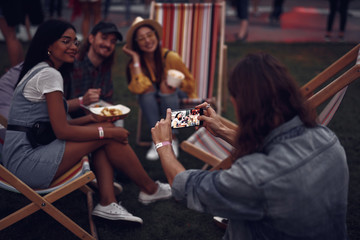 Back view portrait of hipster guy photographing girls with bearded man while they sitting on folding chairs. Focus on phone display with picture