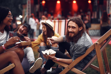 Portrait of cheerful hipster friends sitting on folding chairs and having fun. Focus on smiling bearded guy with food and lovely girl in glasses