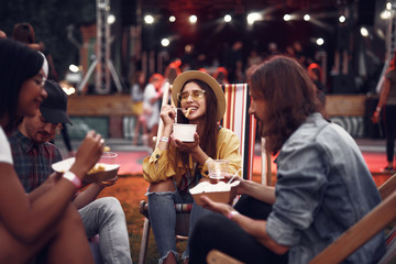 Portrait of cheerful young lady in hat sitting on folding chair and eating french fries while handsome bearded guys holding beer. Stage and crowd on blurred background