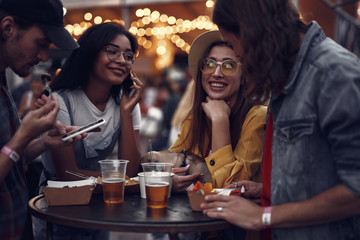 Portrait of young people standing at the table, chatting and using smartphones. Smiling girl in hat holding cute pug dog
