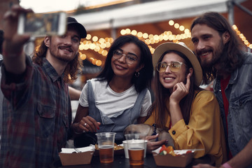 Portrait it of young people taking photo with smartphone while standing at the table in outdoor cafe. Smiling girl in hat holding cute pug dog