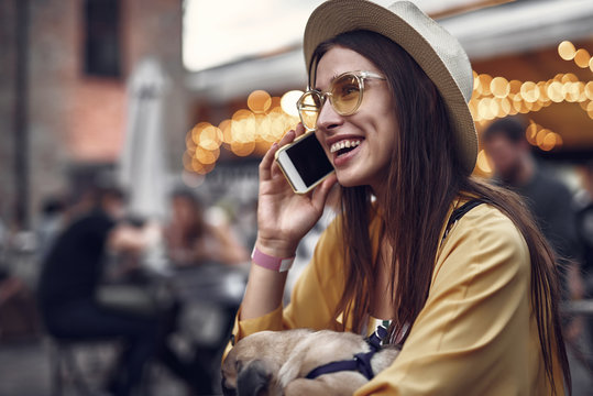 Side view portrait of hipster young lady in hat enjoying phone conversation while holding little pug dog. She is looking away and smiling