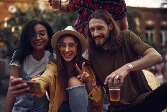 Portrait of stylish hipster friends sitting on car and taking picture with smartphone. Lovely girl in hat showing victory sign while smiling bearded guy holding beer