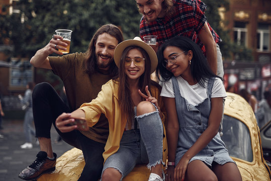Portrait of stylish young people sitting on car and taking picture with smartphone. Smiling bearded guys holding cups with cold beer