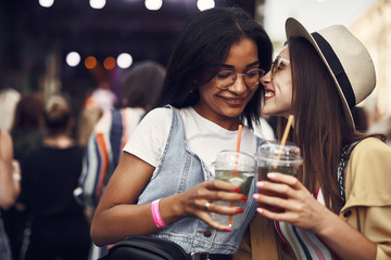 This is secret. Portrait of young smiling lady in hat whispering something to friend. Cheerful women holding drinks