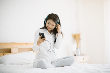 Woman listening music in headphones while sitting on bed in room