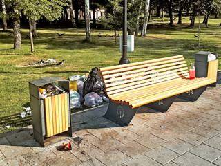 wooden bench and trash cans at picnic area in recreation zone forest city park with full of waste around and crow family looking for food on grass nature outdoor lifestyle ecology background scene
