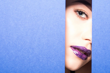 Young beautiful woman with a bright make-up and violet lips looks through a hole in violet paper. Cosmetics, cosmetics, beauty salon, makeup artist, lip gloss, beautiful teeth, sales.