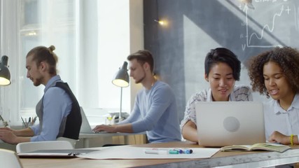Tracking shot of two multiethnic female office workers sitting at desk, using laptop computer together, talking and smiling while other employees are working in the background
