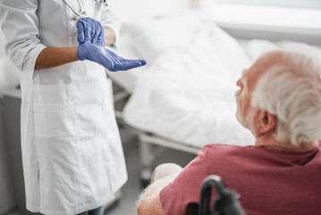 Cropped portrait of female medic in white lab coat preparing for procedure. Old man sitting in wheelchair