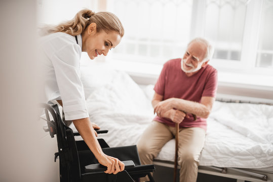 Portrait of smiling young woman in white lab coat preparing wheelchair for bearded gentleman while he sitting on bed. Man holding walking stick and looking at lady with smile